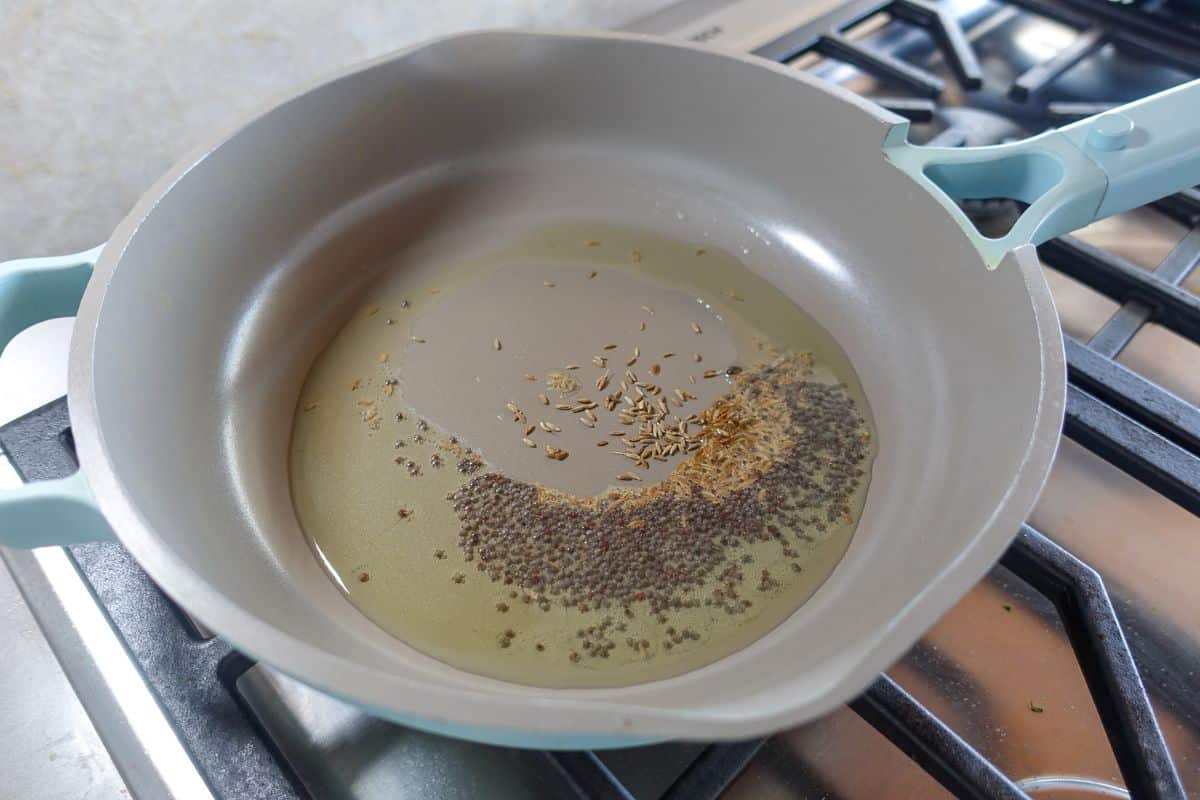 A light blue frying pan on a stove with oil, mustard seeds, and cumin seeds being heated.