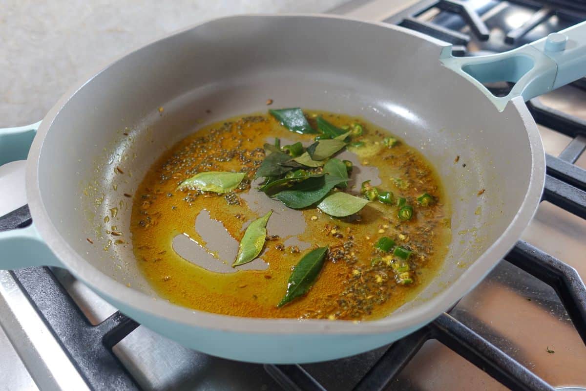A frying pan on a stovetop contains oil, curry leaves, green chilies, and spices sizzling together.