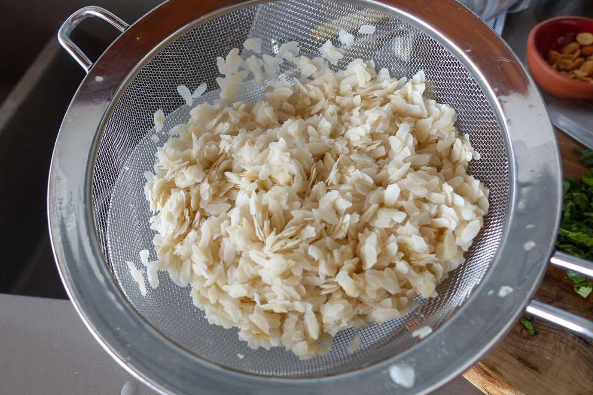 A metal strainer filled with rinsed, flattened rice flakes sits on a kitchen counter next to a cutting board and small bowls.