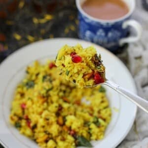 A close-up of a spoonful of aloo poha with pomegranate seeds held above a plate of the same, with a cup of tea in the background.