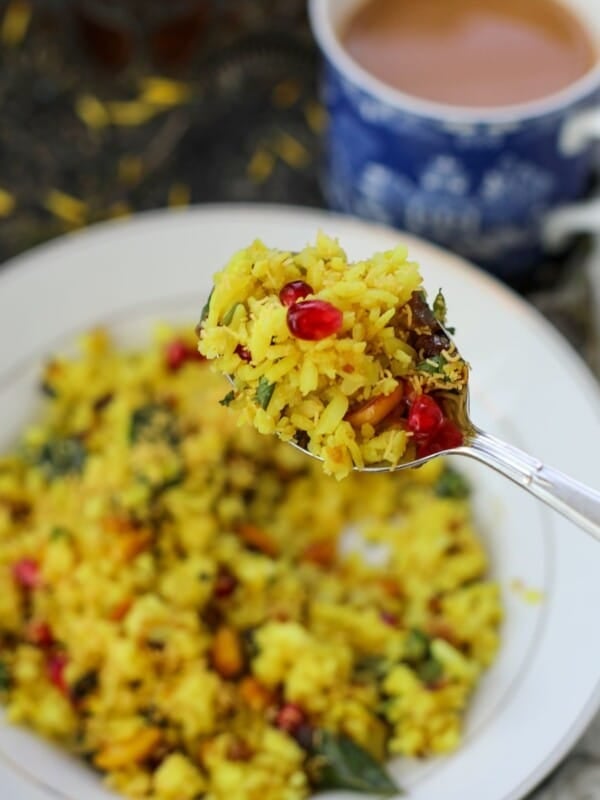 A close-up of a spoonful of aloo poha with pomegranate seeds held above a plate of the same, with a cup of tea in the background.