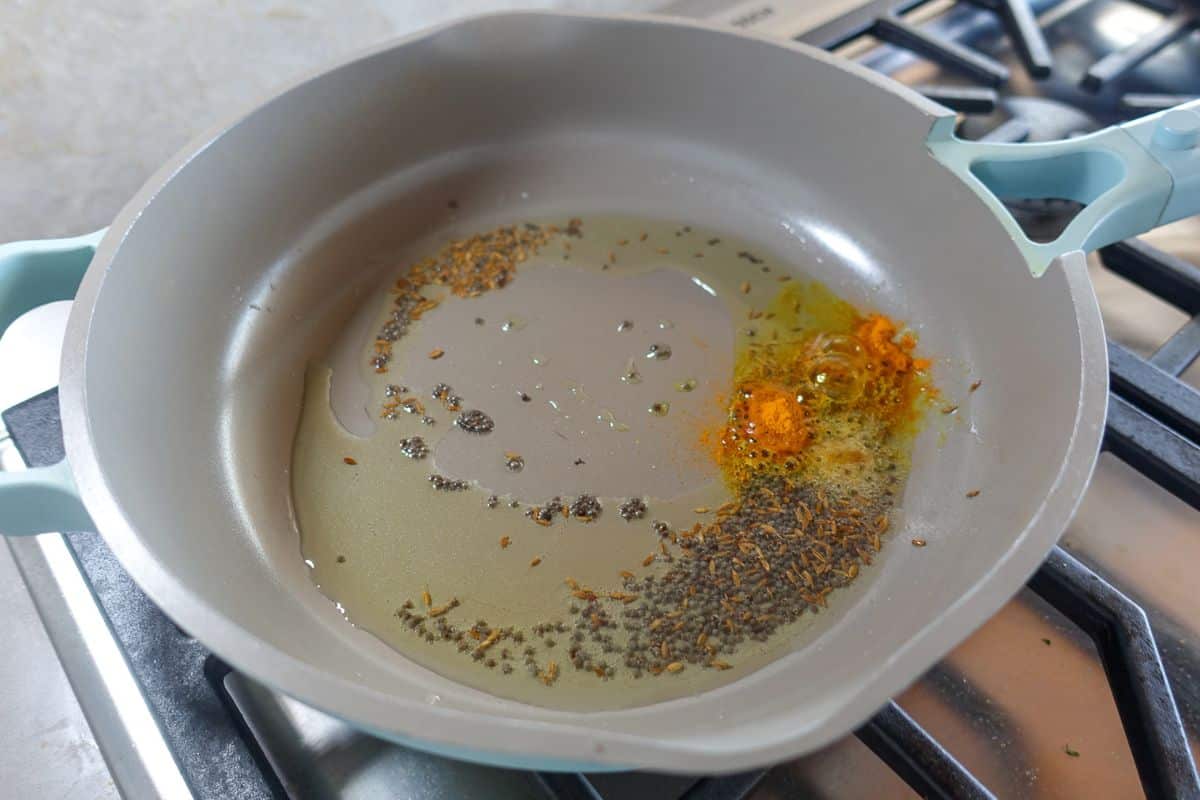 A frying pan on a gas stove contains oil, cumin seeds, black mustard seeds, and turmeric powder being heated.