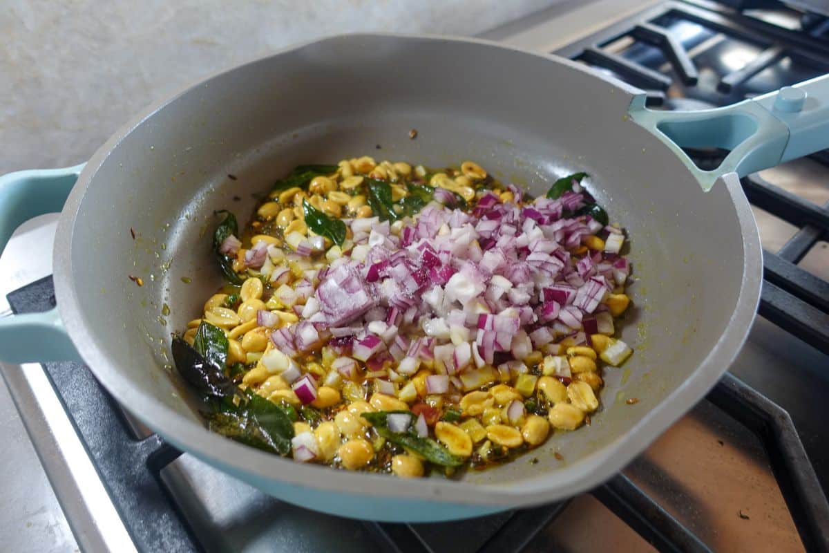 A frying pan on a stove contains chopped red onions, peanuts, curry leaves, and spices being sautéed.