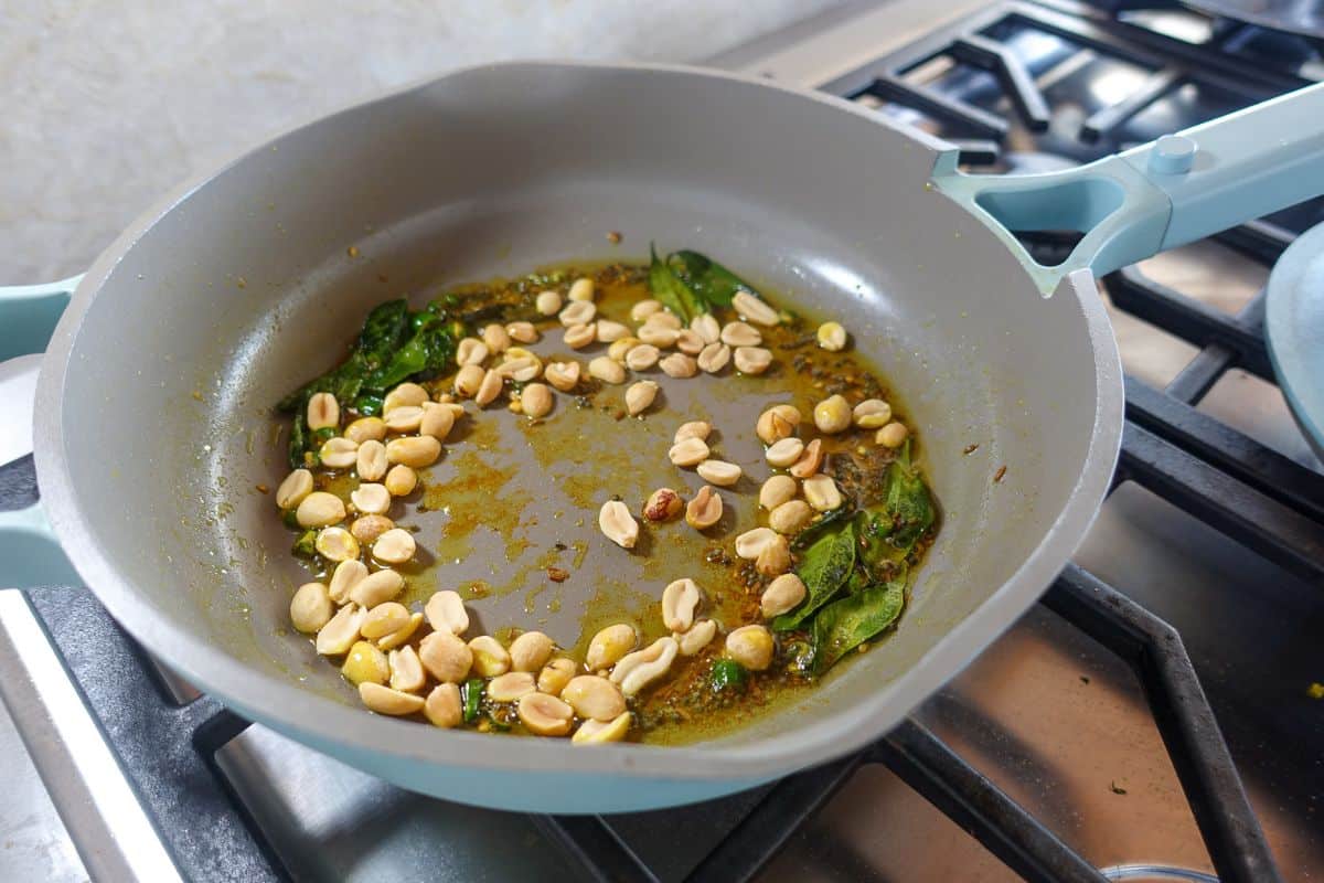 A light blue frying pan on a stove contains peanuts, green curry leaves, and oil being sautéed.