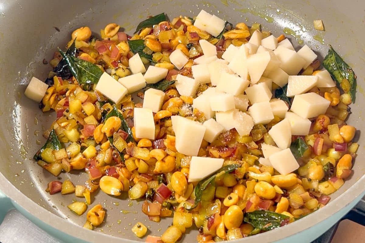 Diced potatoes, peanuts, curry leaves, and chopped onions are being sautéed in a pan with oil and spices.