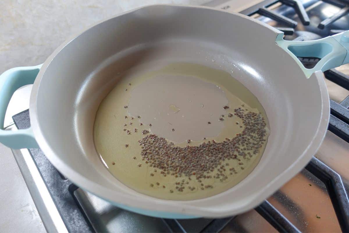 A frying pan with oil and mustard seeds heating on a stovetop burner.