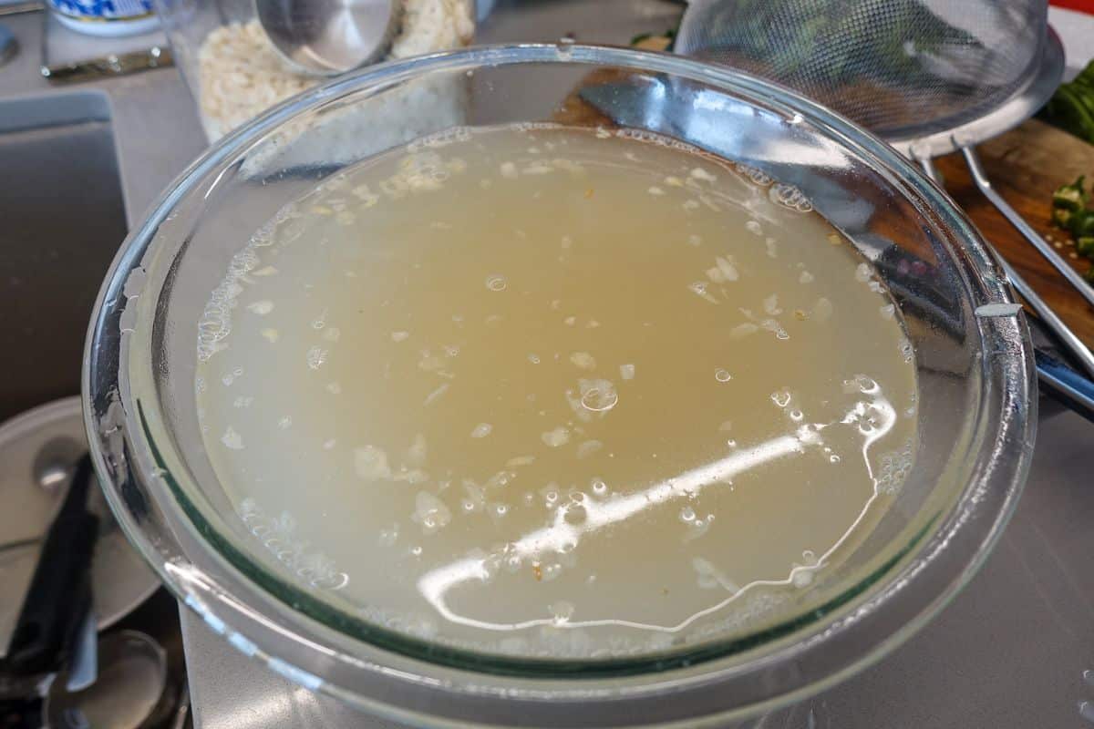 A clear glass bowl filled with cloudy rice water sits on a kitchen counter near utensils and a strainer.