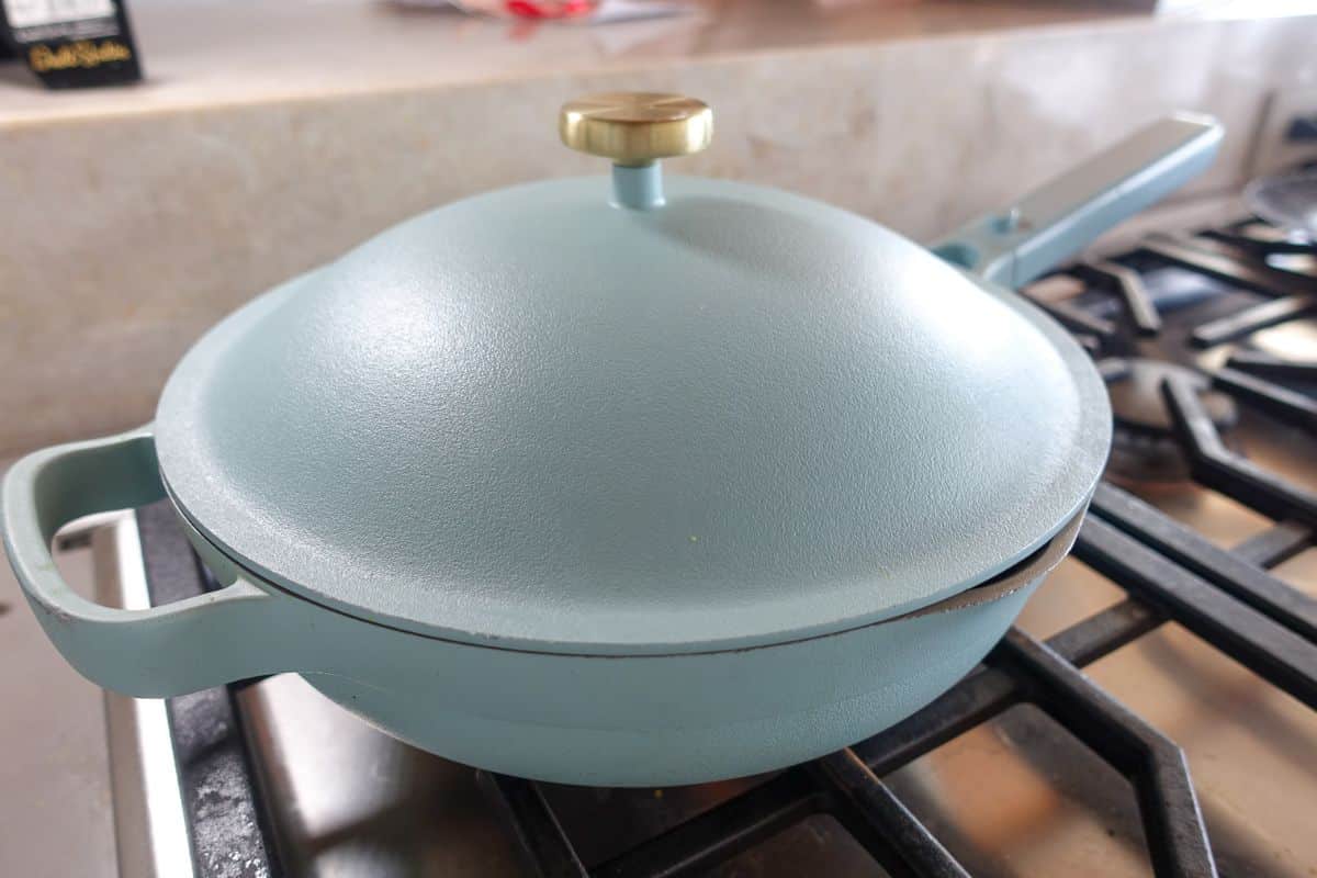 A light blue cast iron pan with a lid and brass knob sits on a gas stove burner in a kitchen setting.