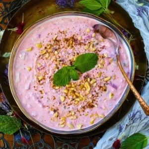 A bowl of pink Beetroot Raita garnished with chopped peanuts, a sprinkle of spices, and fresh mint leaves, with a spoon resting in the bowl.