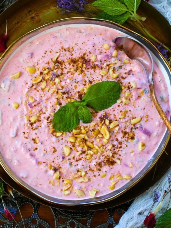 A bowl of pink Beetroot Raita garnished with chopped peanuts, a sprinkle of spices, and fresh mint leaves, with a spoon resting in the bowl.