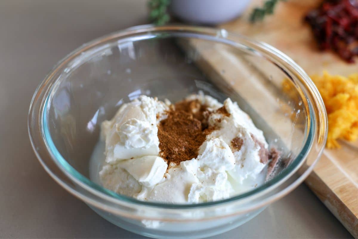 A clear glass bowl contains a mixture of Greek Yogurt with roasted cumin, red chili powder, and pink salt, set on a countertop near a cutting board with orange zest and dried chili peppers.