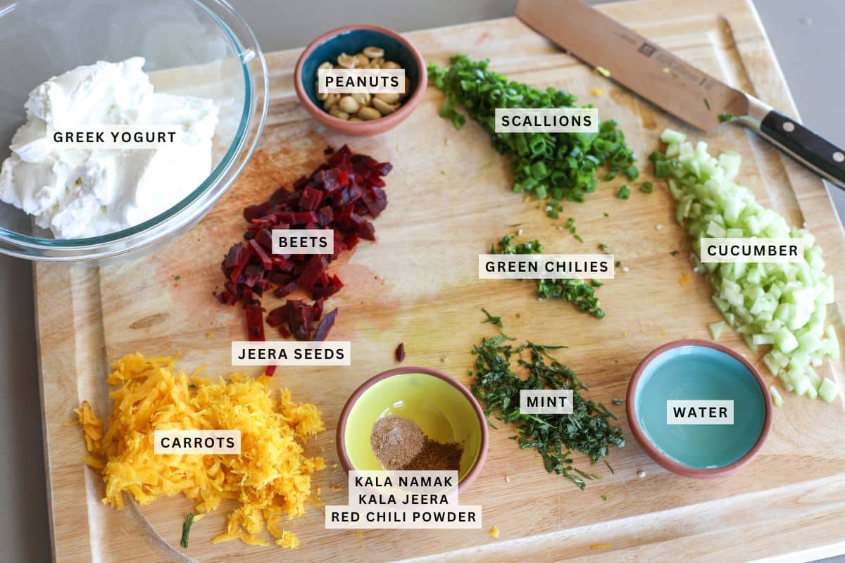 Chopped beets, carrots, scallions, cucumber, green chilies, mint, peanuts, spices, water, and Greek yogurt labeled on a wooden cutting board with a knife.