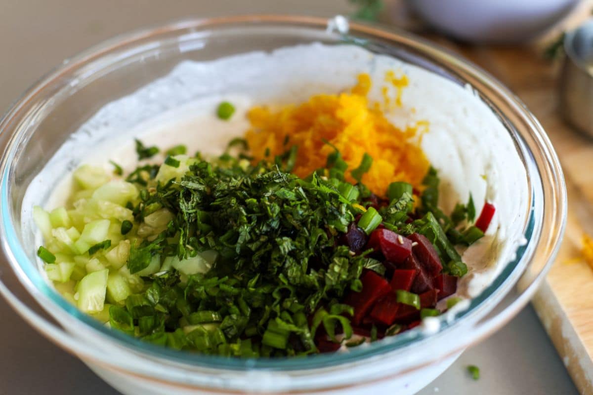 A glass bowl containing chopped green onions, cucumbers, beets, shredded carrots, and mint on top of a creamy white base.