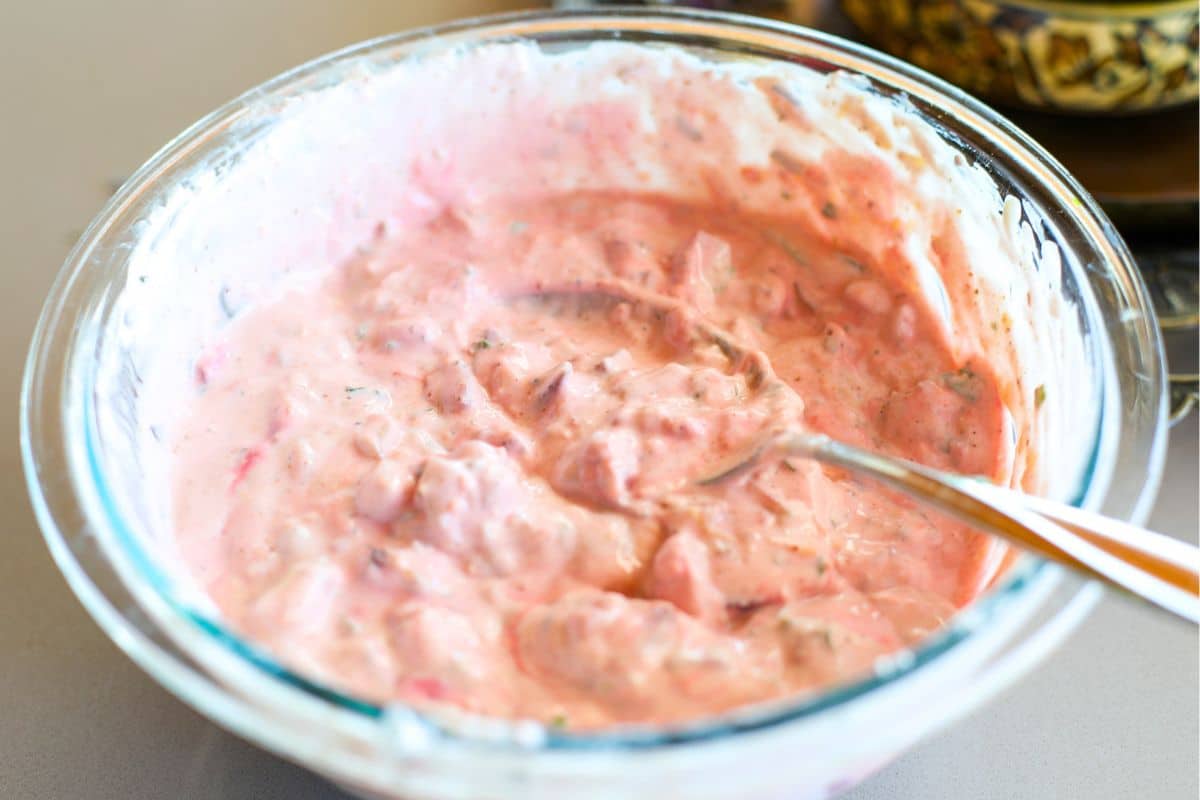 A glass bowl filled with Beetroot raiata containing chopped vegetables, with a spoon resting inside the mixture.