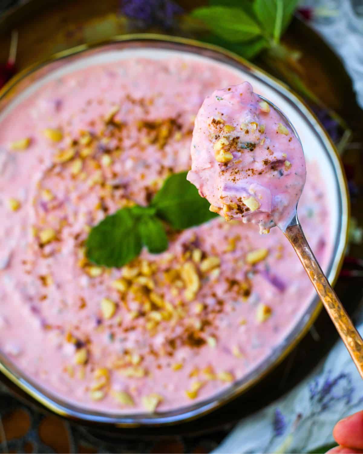 A bowl of creamy pink Beetroot Raita topped with chopped peanuts, roasted cumin, and mint leaves, with a spoonful of the raita held above the bowl.