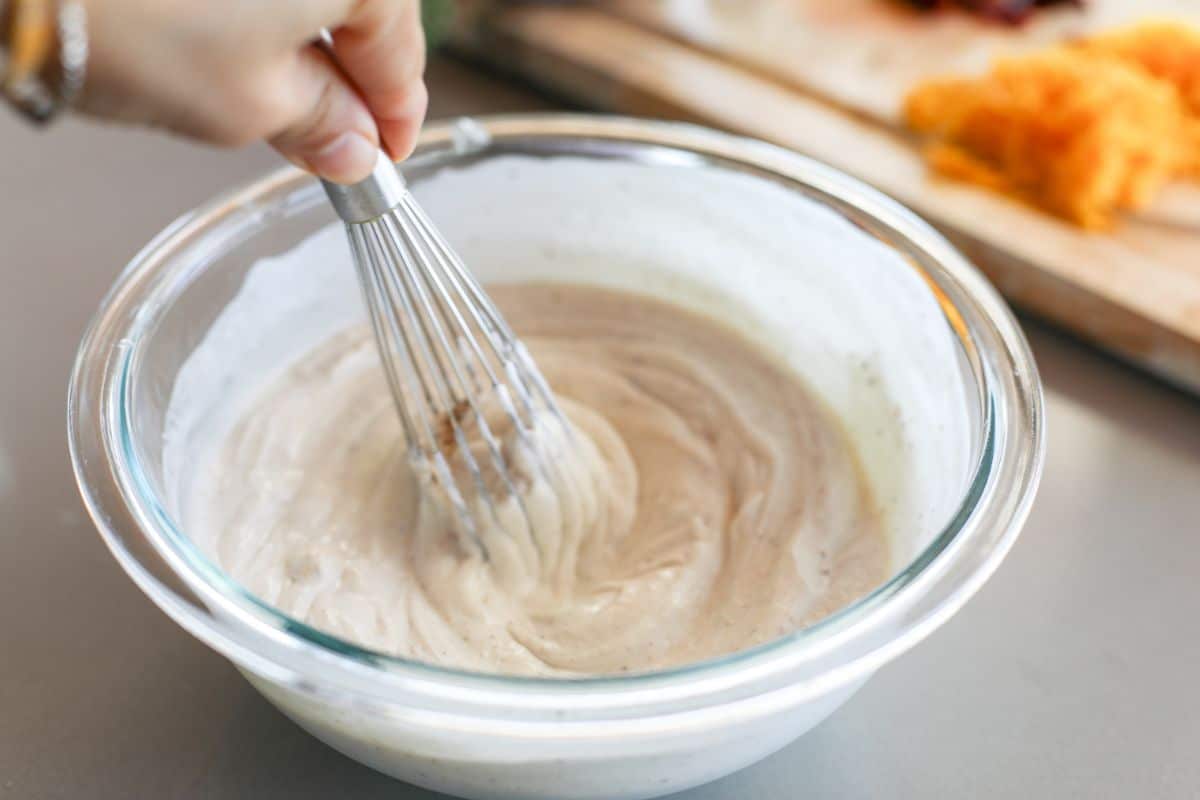A hand using a whisk to mix batter in a clear glass bowl on a kitchen counter.