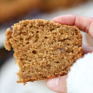 A hand holding a thick slice of moist eggless banana bread with jaggery and ghee with a rough texture and a partially bitten corner.