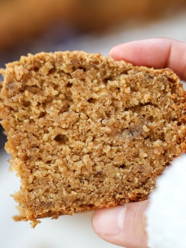 A hand holding a thick slice of moist eggless banana bread with jaggery and ghee with a rough texture and a partially bitten corner.