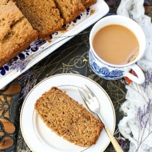 A slice of banana bread on a plate with a fork, next to a cup of coffee with milk and a platter holding more slices of banana bread.