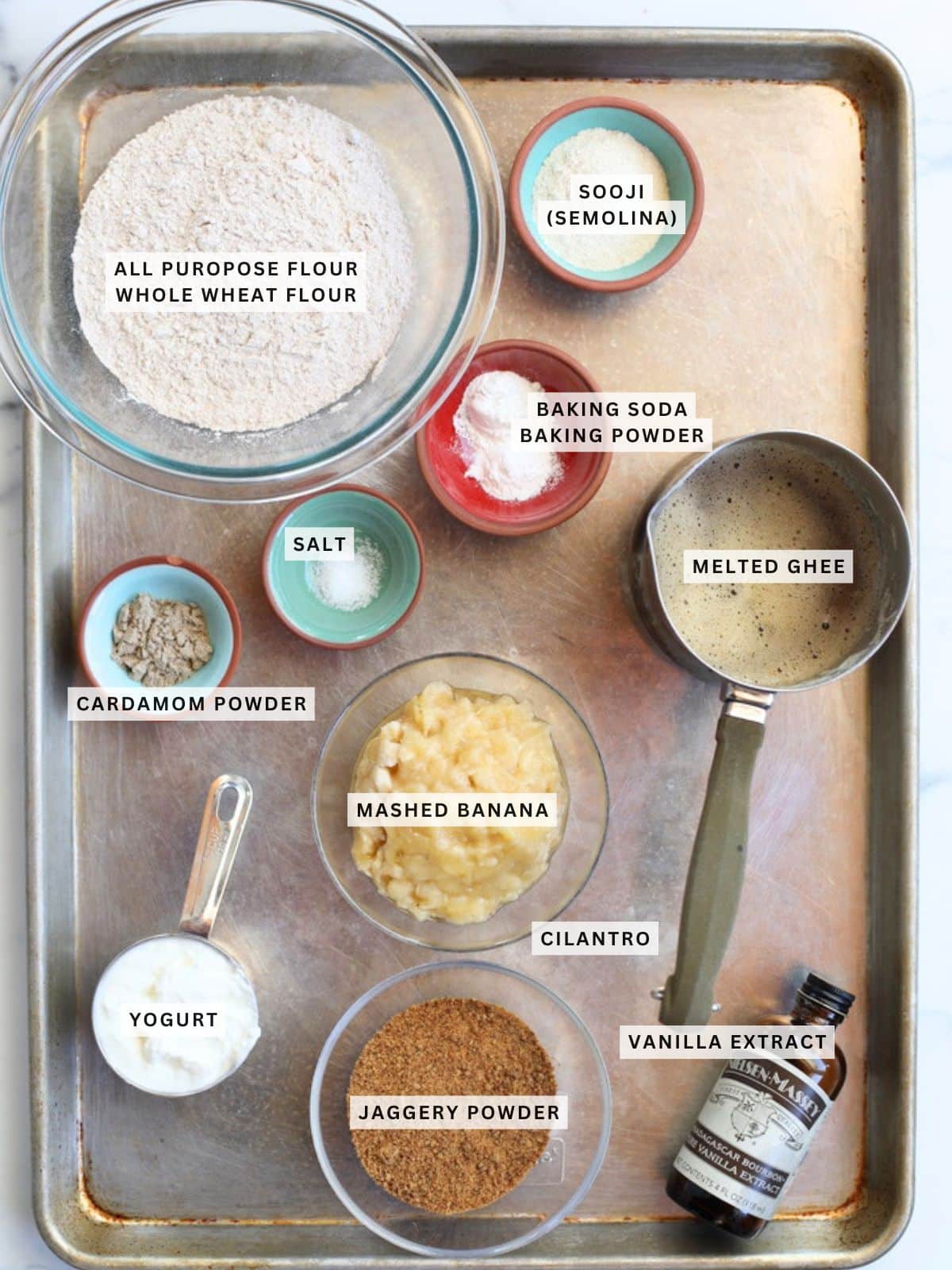 A tray holds bowls and cups containing labeled ingredients for baking, including flours, semolina, baking powder, baking soda, salt, cardamom, ghee, banana, yogurt, jaggery, and vanilla extract.
