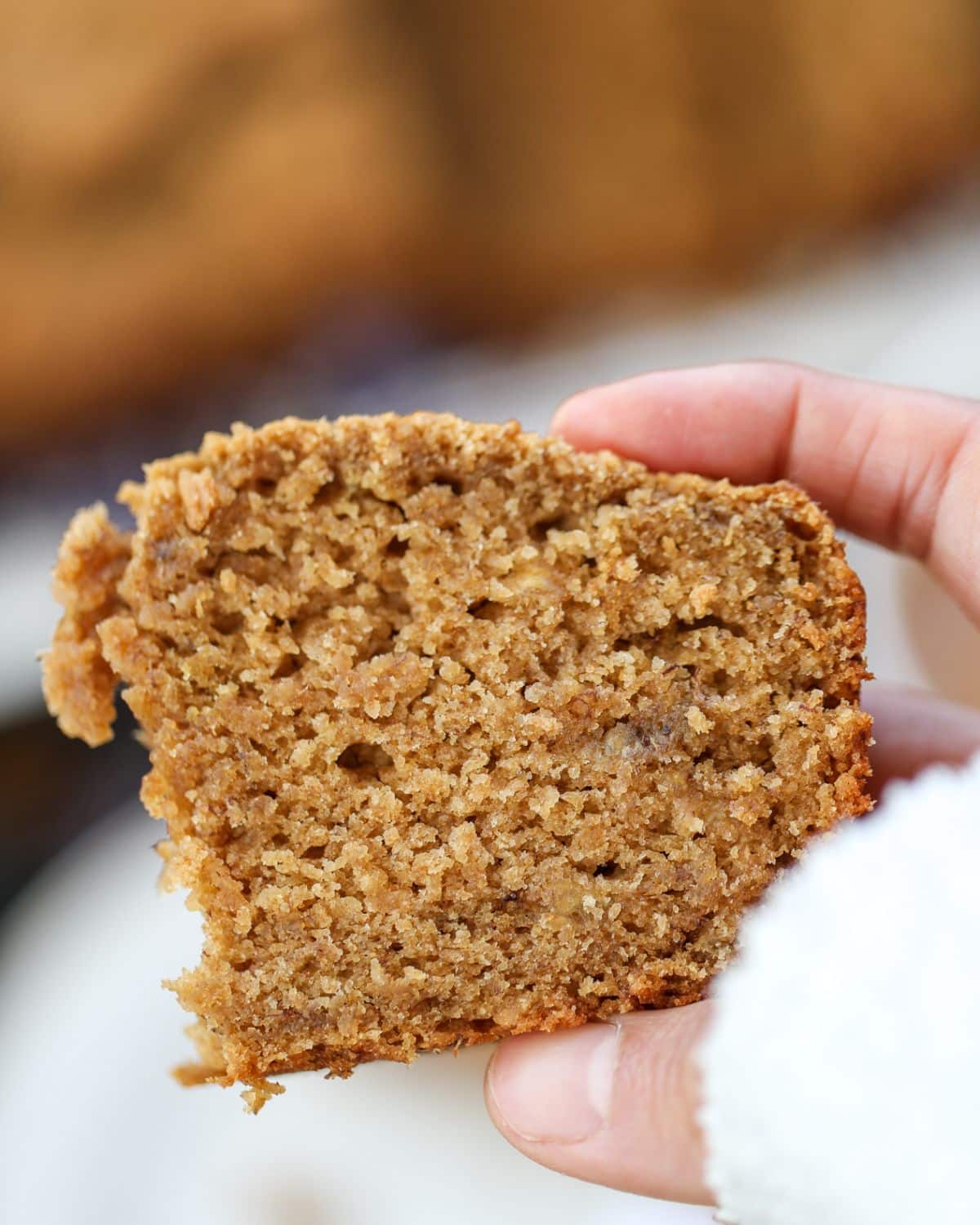 A hand holding a thick slice of moist eggless banana bread with jaggery and ghee with a rough texture and a partially bitten corner.