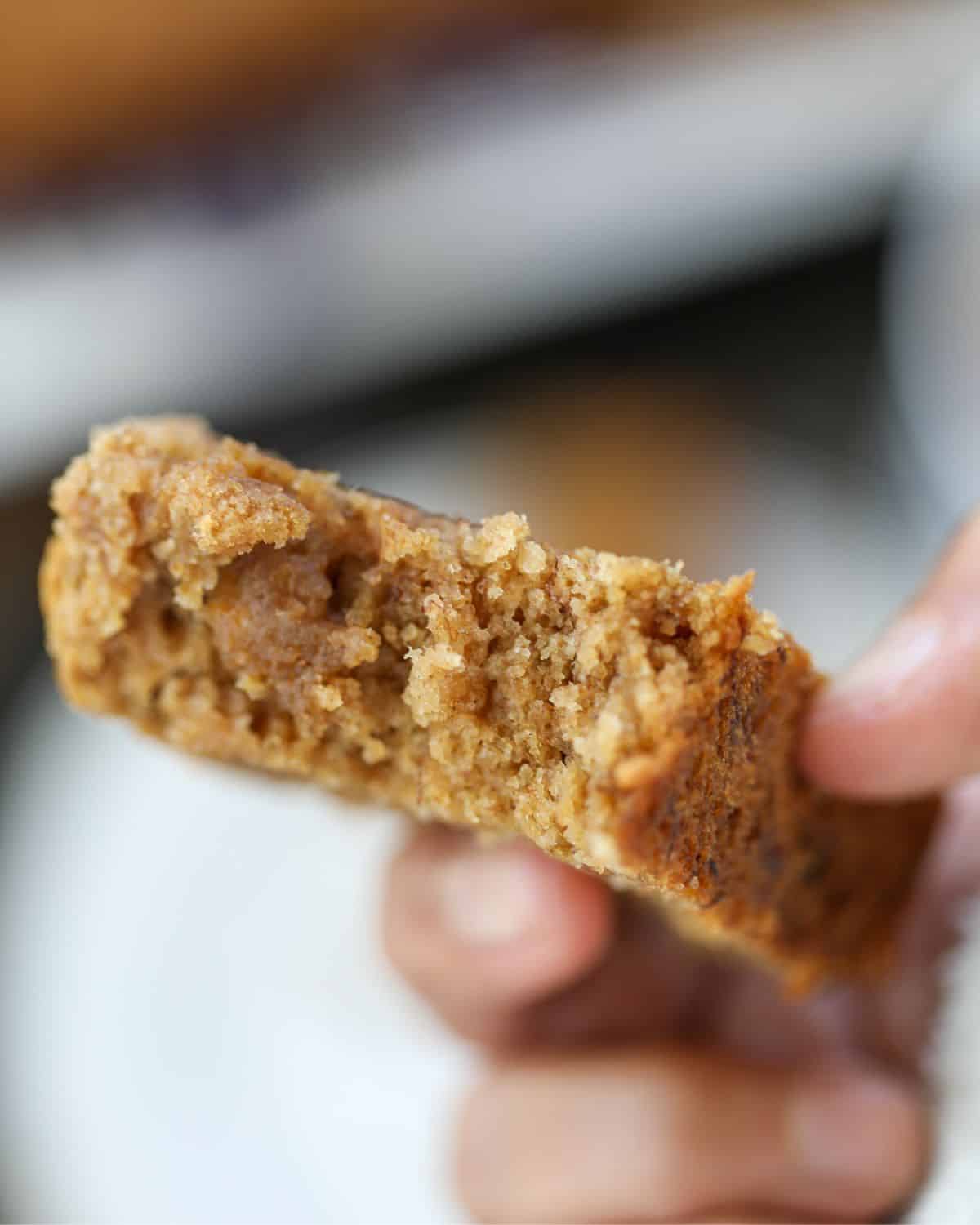 A hand holding a thick slice of moist eggless banana bread with jaggery and ghee with a rough texture and a partially bitten corner.