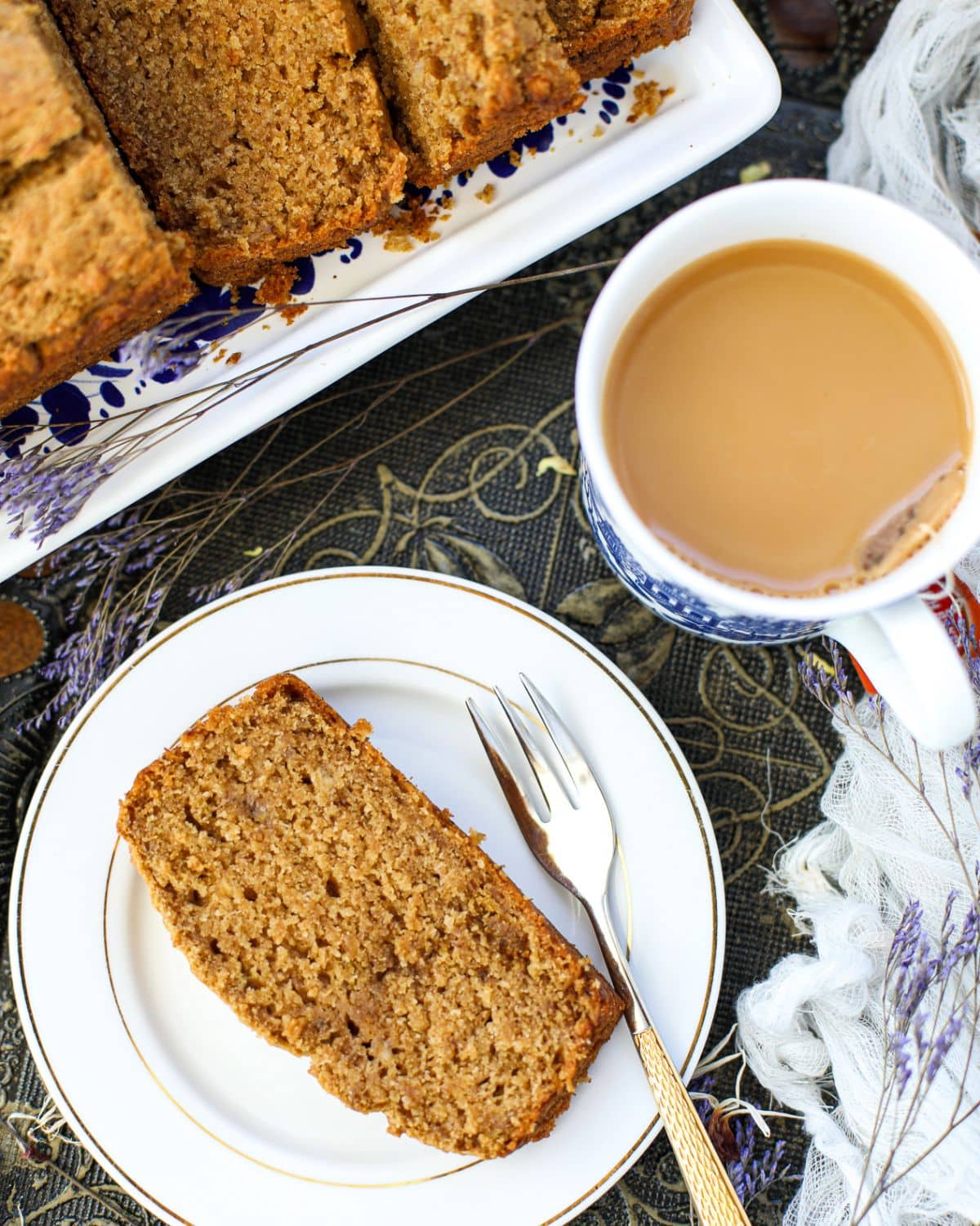 A slice of eggless banana bread with jaggery and ghee on a white plate with a fork, next to a cup of coffee with milk and a platter of sliced bread.