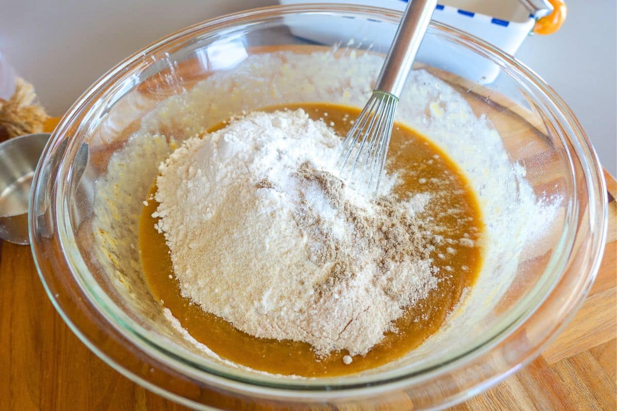A glass bowl containing flour, spices, and wet ingredients being mixed with a whisk, set on a wooden surface.