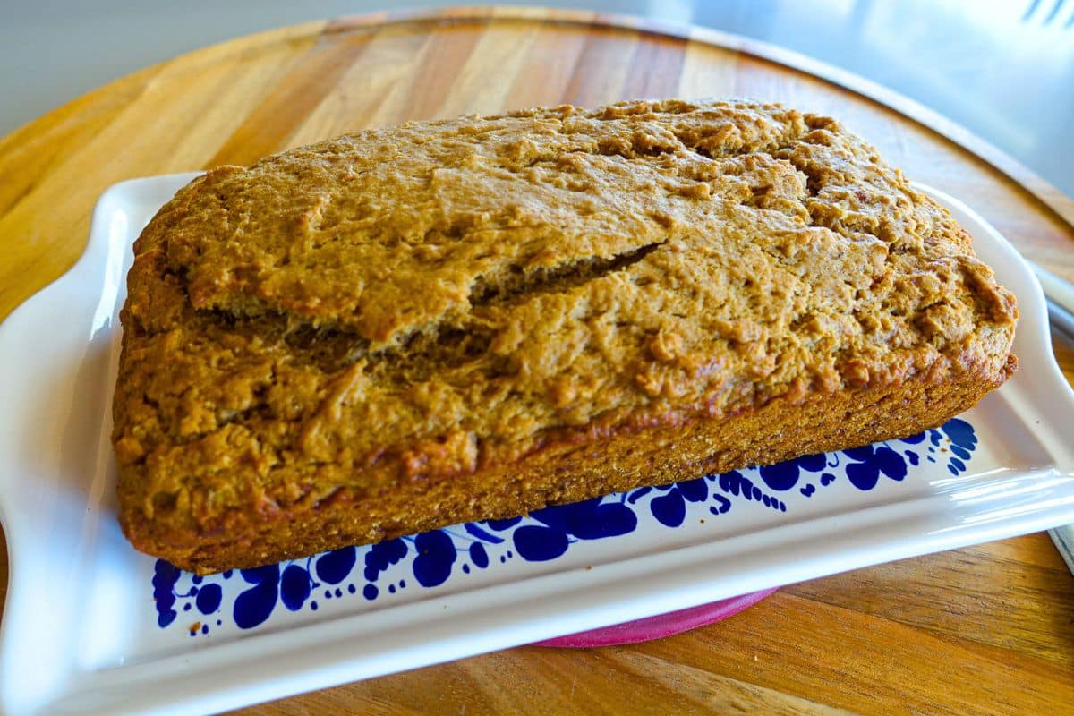 A loaf of homemade eggless banana bread sits on a white and blue ceramic tray atop a wooden surface.
