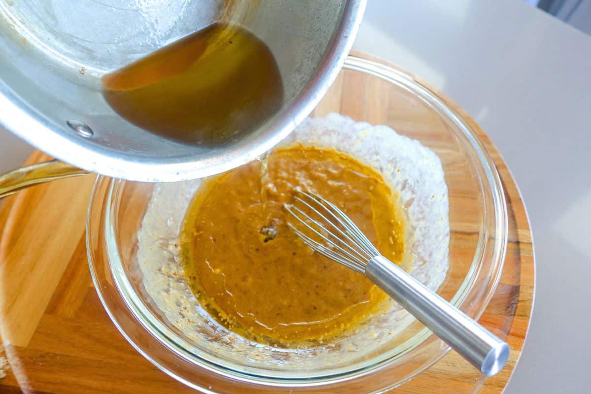 A saucepan pours melted ghee into a glass bowl containing a brown mixture with a whisk on a wooden board.