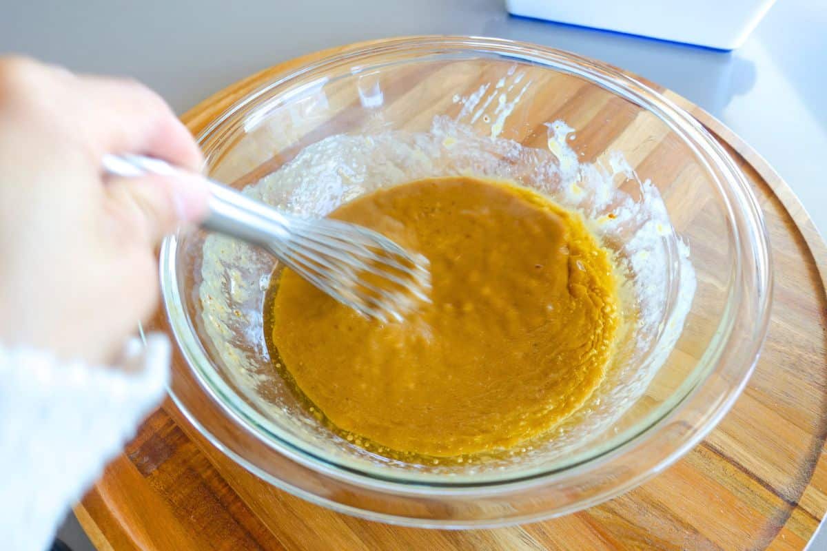 A person uses a whisk to mix a thick, yellow-brown batter in a clear glass bowl placed on a wooden surface.