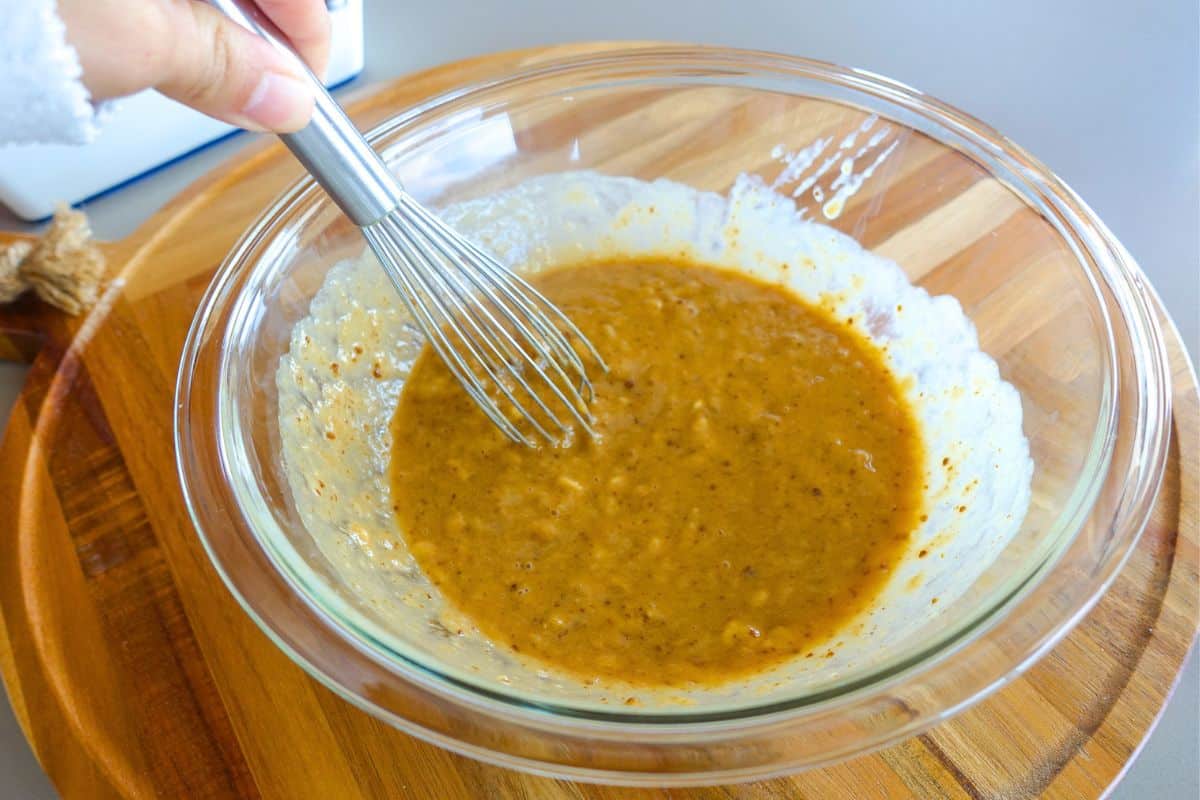 A hand holds a whisk mixing a thick, brown batter in a clear glass bowl on a wooden surface.
