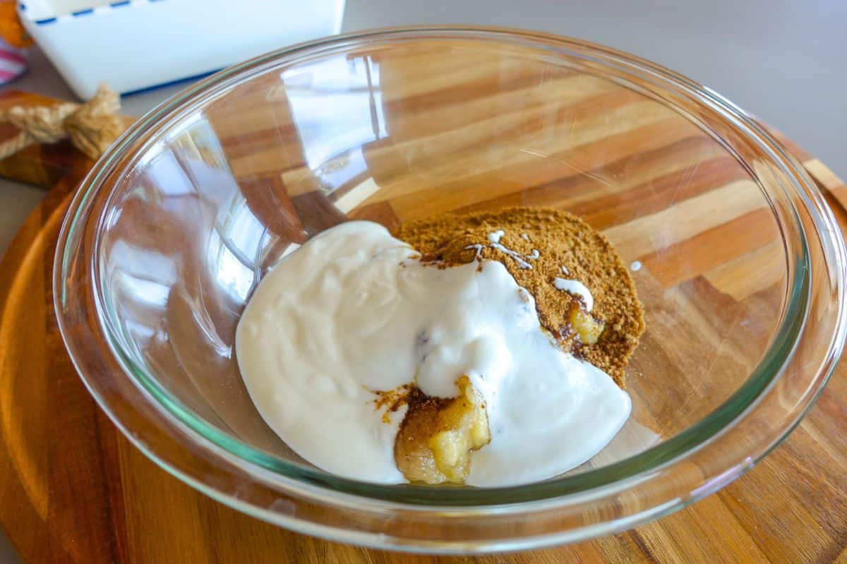 A glass bowl on a wooden surface containing jaggery, yogurt, mashed banana, and vanilla extract.