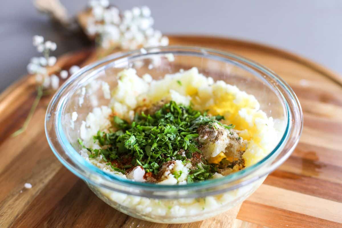 A glass bowl of mashed potatoes topped with chopped herbs and spices sits on a wooden board.