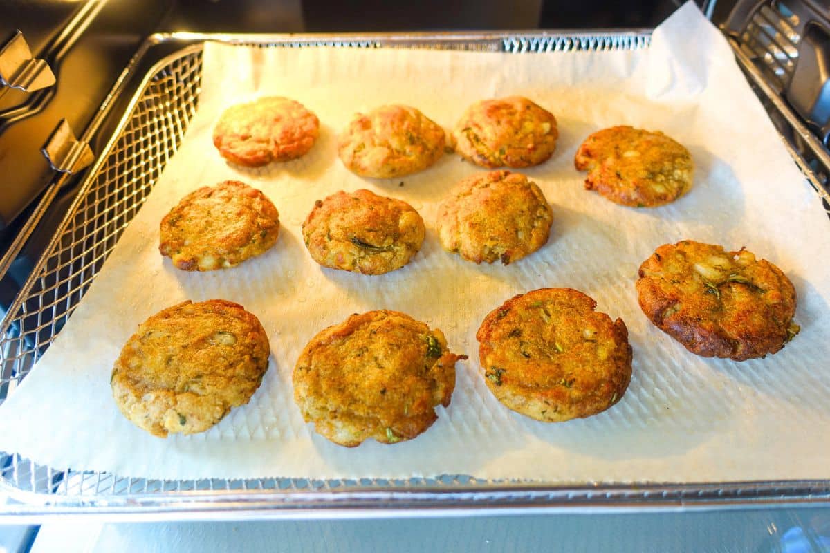 Ten round, golden-brown potato patties are arranged on parchment paper in an air fryer basket, positioned inside an oven.