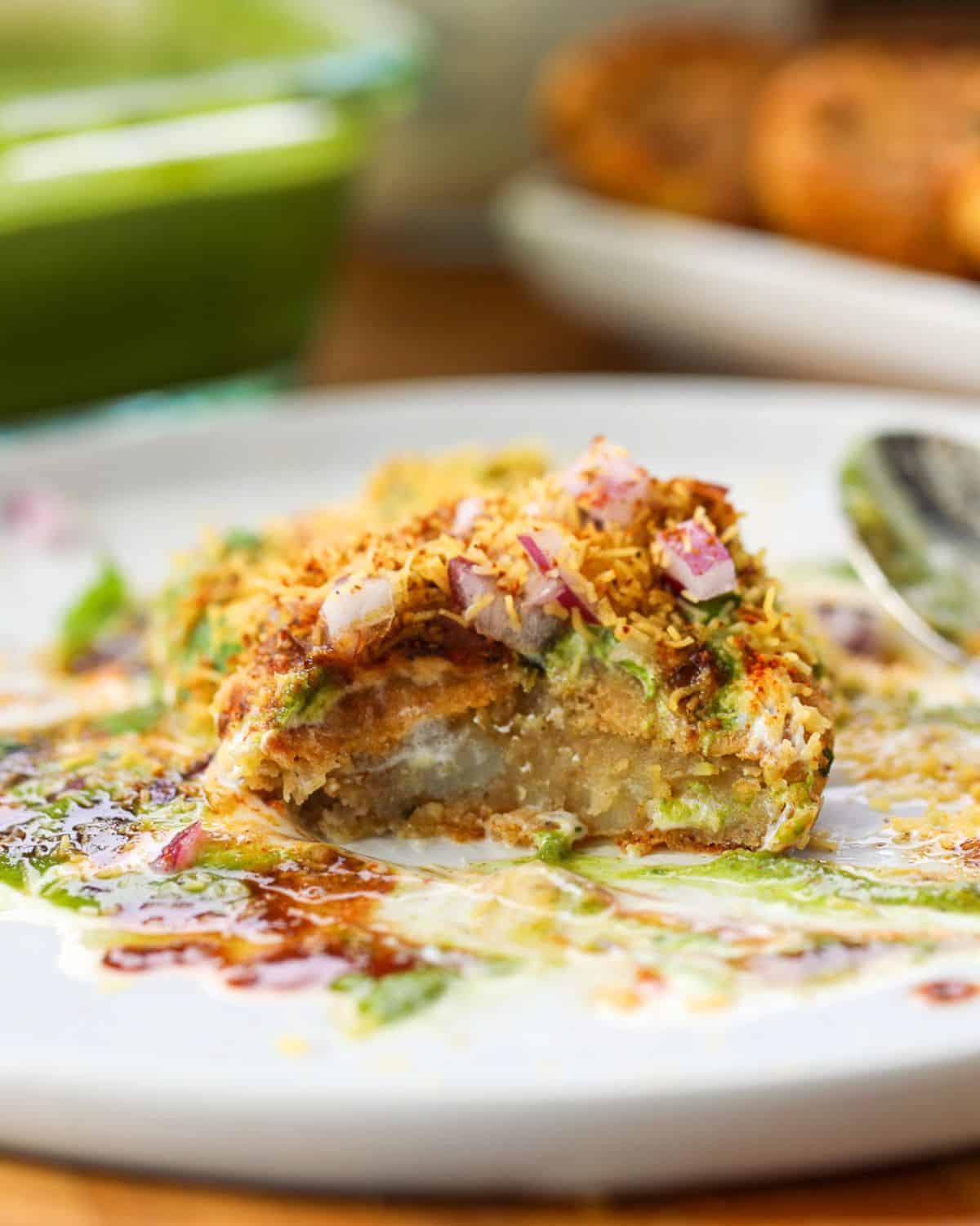A half-eaten aloo tikki chaat on a white plate, topped with sev, chopped onions, and chutneys, with a spoon beside it.
