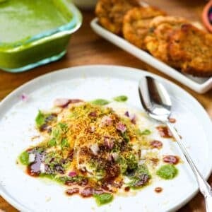 A plate of aloo tikki chaat garnished with chutneys, onions, and sev, with a spoon beside it; green chutney and fried snacks in the background.