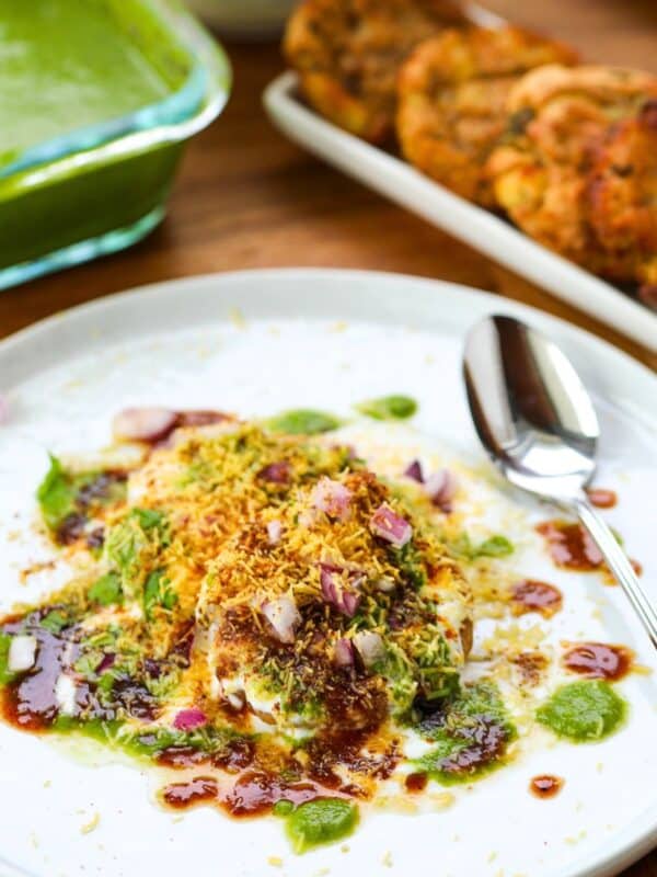 A plate of aloo tikki chaat garnished with chutneys, onions, and sev, with a spoon beside it; green chutney and fried snacks in the background.