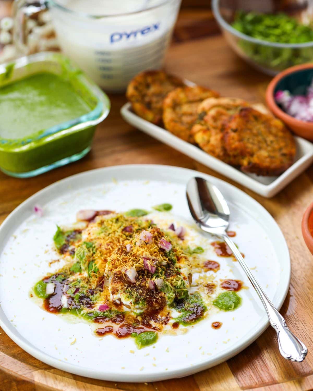 A plated aloo tikki chaat dish garnished with chutneys and onions, surrounded by a spoon, green chutney, measuring cup, fried patties, and chopped herbs on a wooden table.