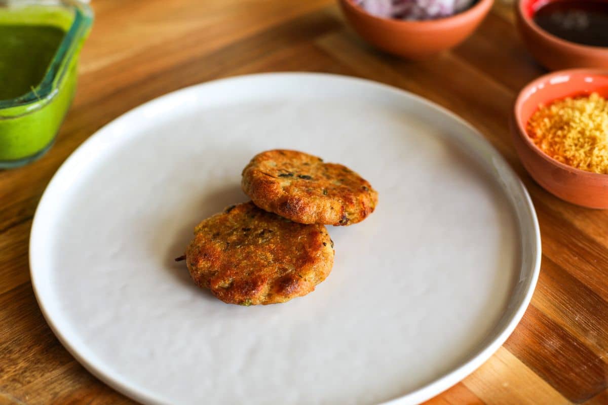 Two golden-brown potato patties are placed on a plain white plate, with small bowls of chutney, onions, and sev in the background on a wooden table.