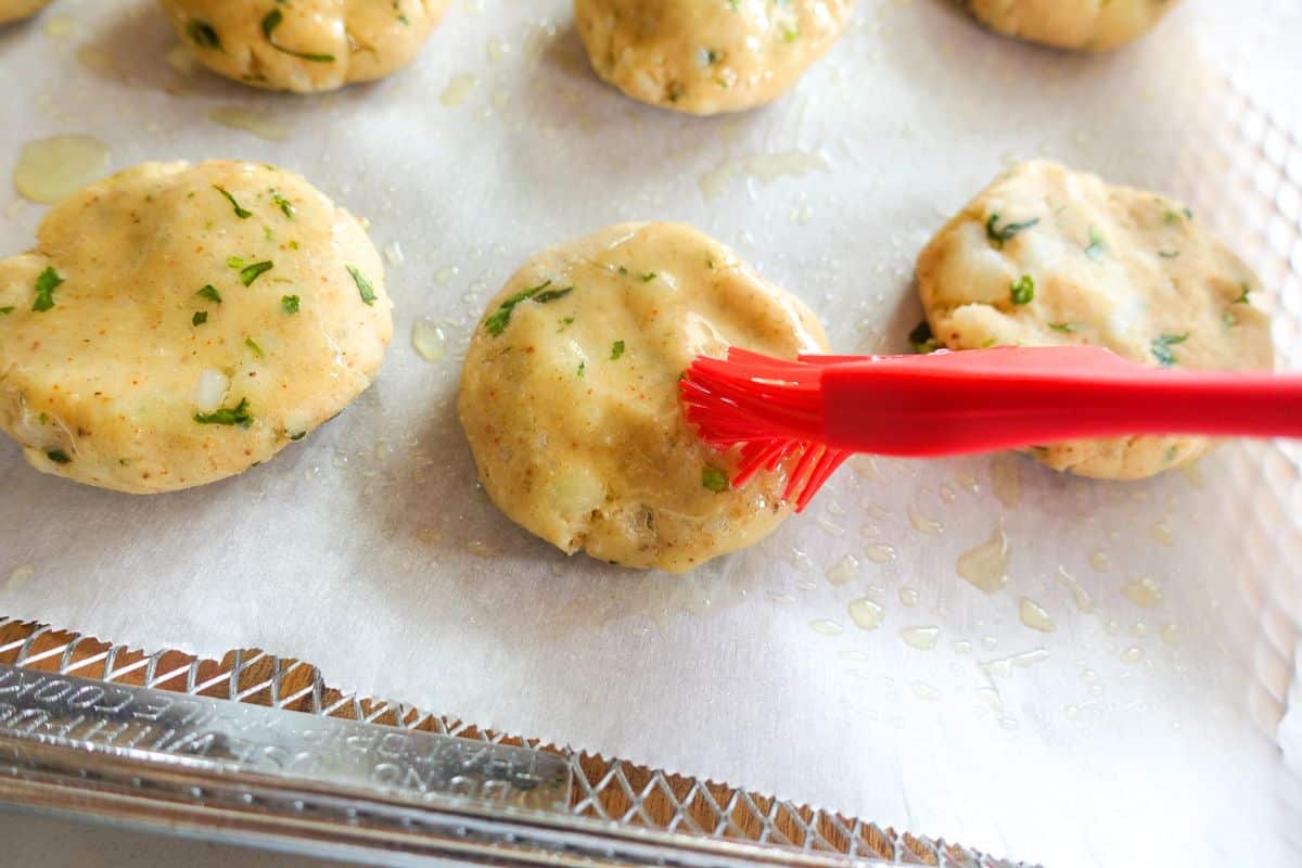 A red brush applies oil to uncooked, round potato patty pieces with herbs, arranged on a parchment-lined baking sheet.