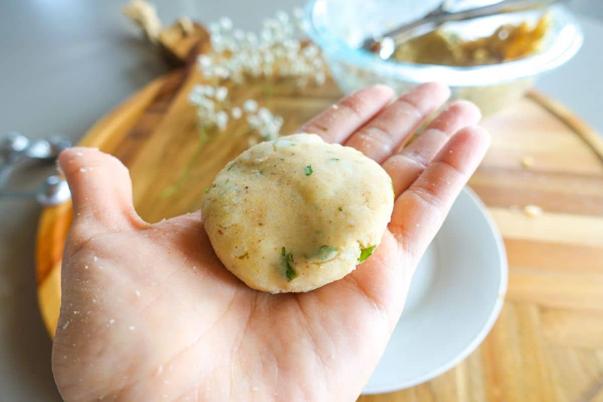 A hand holding a round, uncooked potato patty with green herbs, with a mixing bowl, spoon, and cutting board in the background.