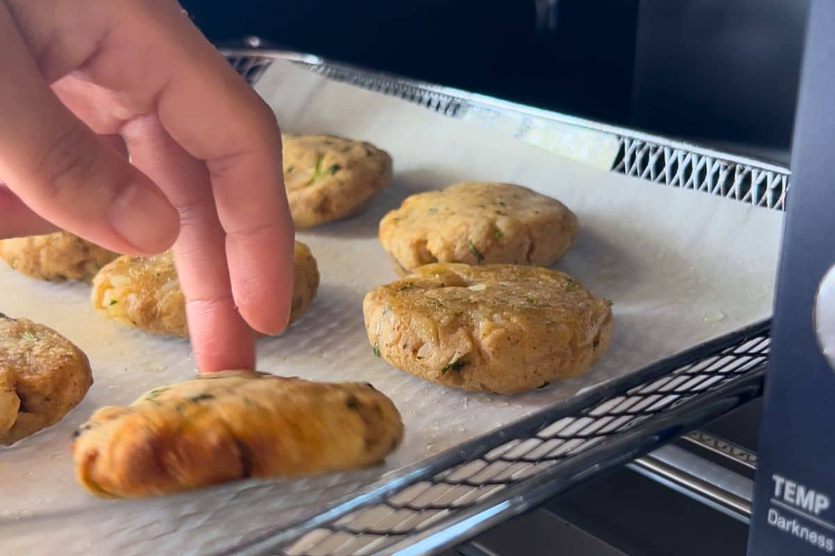 A hand touches a baked potato patty on a parchment-lined tray with several other patties, inside an oven.