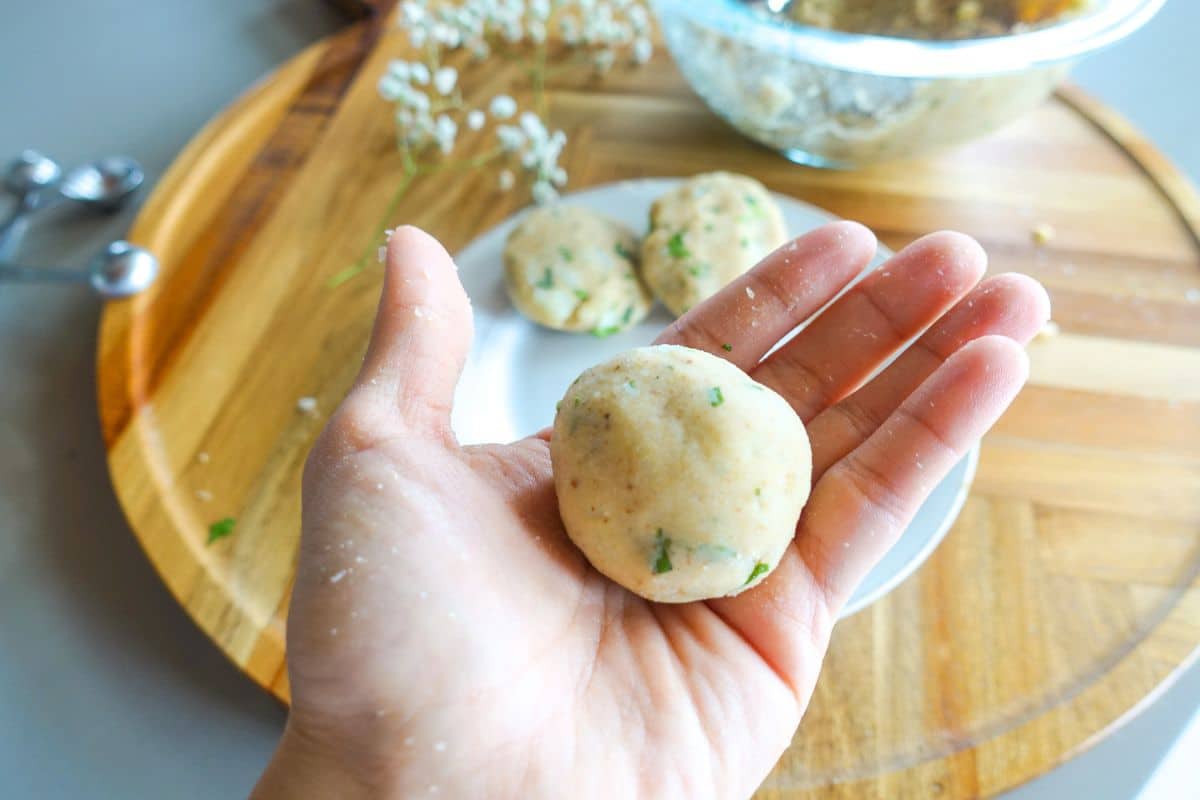 A hand holds a round, uncooked potato ball with herbs; more dough balls and a bowl are on a wooden tray in the background.