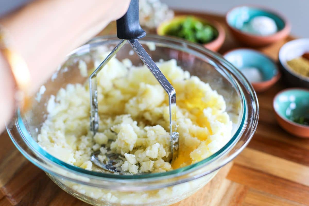 A hand uses a potato masher to mash boiled potatoes in a glass bowl, with small bowls of ingredients in the background on a wooden surface.