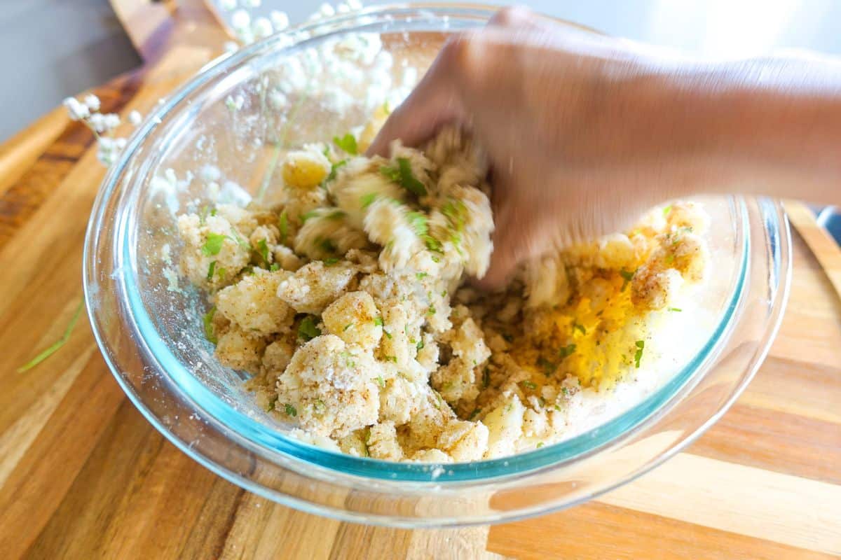 A hand mixes bread cubes, herbs, and other ingredients in a large glass bowl on a wooden surface.