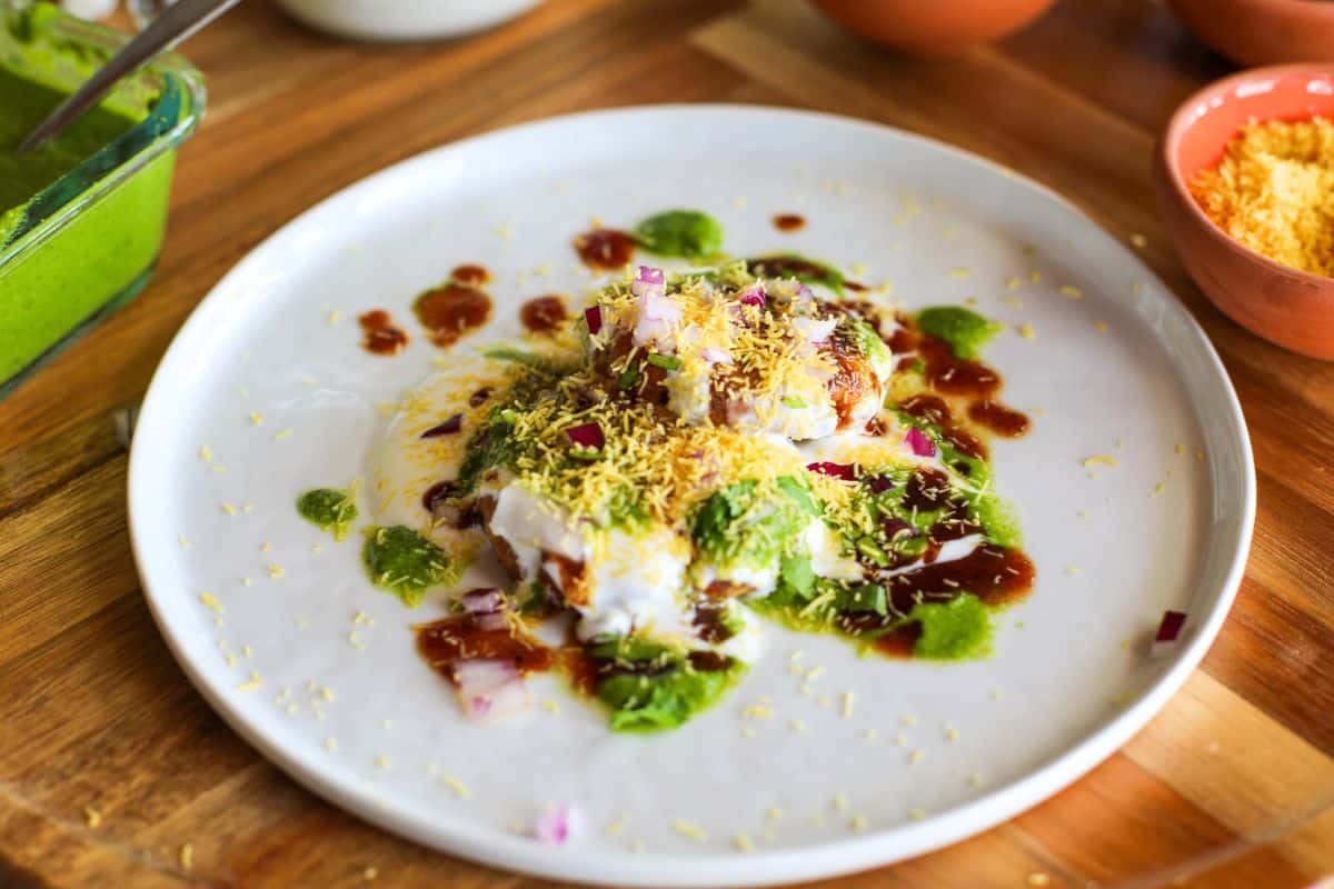 A plate of aloo tikki chaat topped with yogurt, green and tamarind chutneys, chopped onions, and sev on a wooden table.