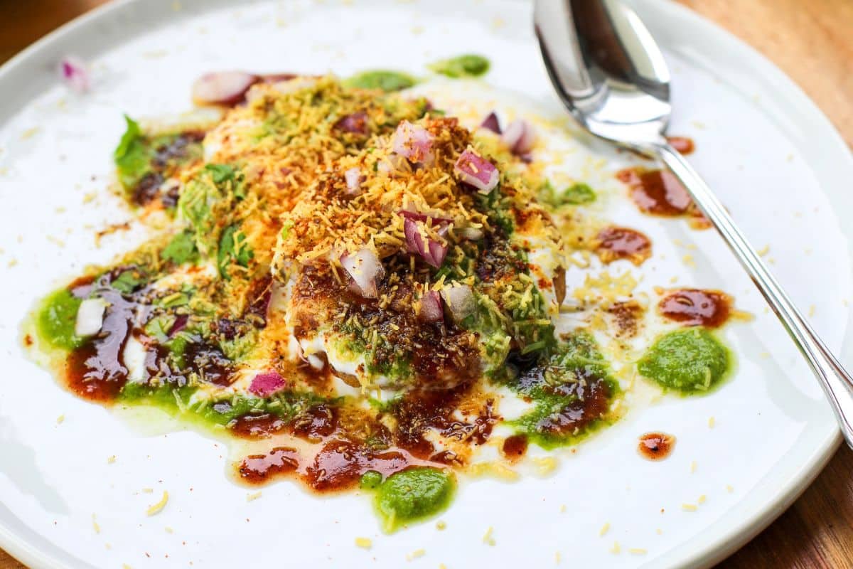 A plate of Indian aloo tikki chaat topped with yogurt, chutneys, sev, and chopped onions, with a spoon on the side.