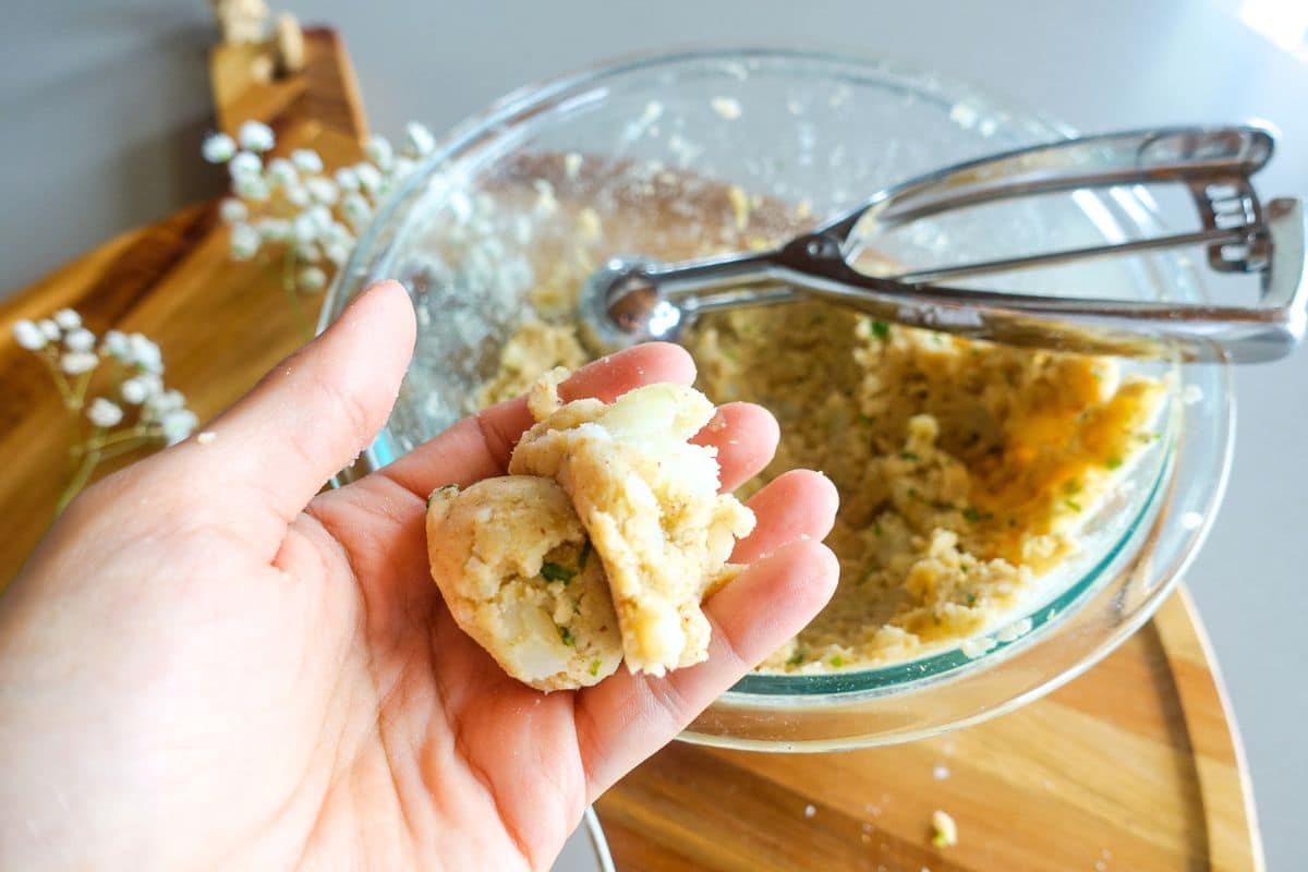 A hand holds a portion of unbaked potato with herbs in front of a bowl of dough and a metal scoop on a wooden surface.