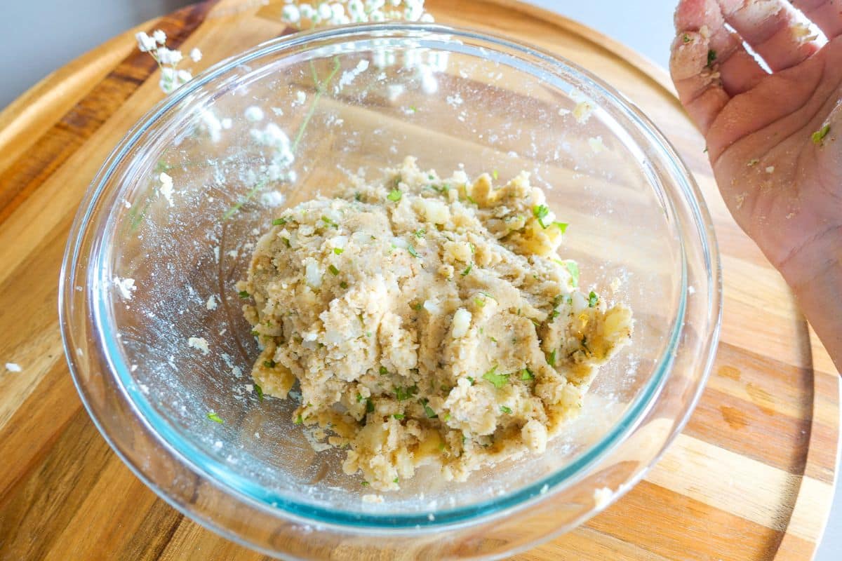 A glass bowl with a mixture of dough and herbs sits on a wooden surface; a hand with dough residue is visible on the right.